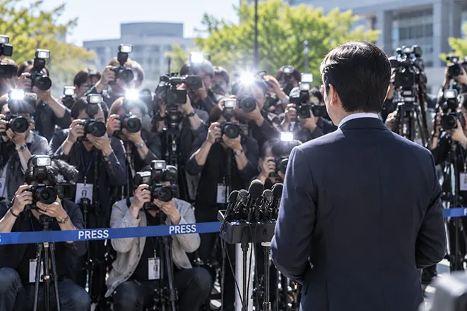 밝은 낮 야외 기자회견에서 여러 사진기자가 카메라 플래시를 사용해 인물을 촬영하는 모습